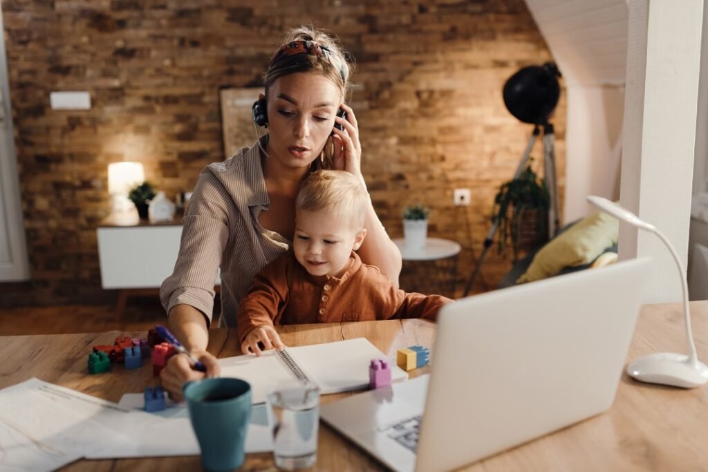 A stay=at-home mom working on her laptop on her online business and has her kid playing on the desk.